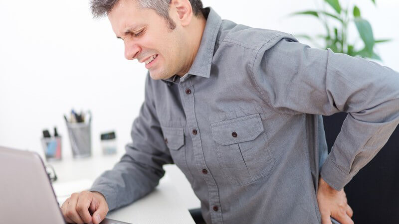 Mann in grauem Hemd sitzt am Laptop und fasst sich vor Rückenschmerzen an den Rücken