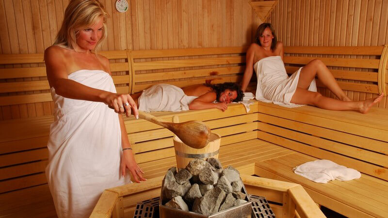 Drei Freundinnen in Sauna, eine macht Aufguss auf heißen Steinen