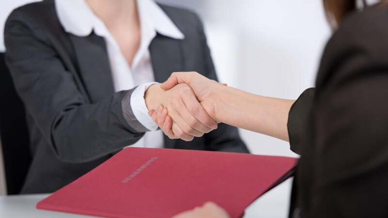 Handschlag zwischen zwei Businessfrauen, die vordere Frau mit Bewerbungsmappe in der linken Hand
