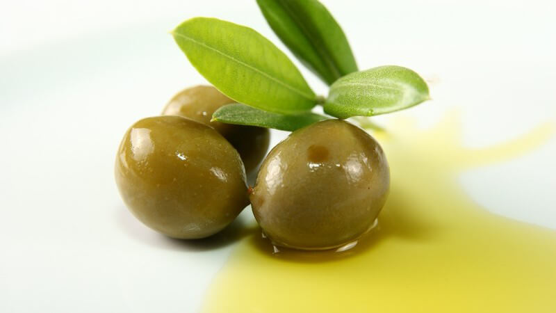 Drei grüne Oliven mit Blättern liegen im gelben Olivenöl