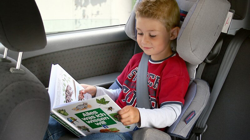 Kleiner Junge angeschnallt im Kindersitz im Auto schaut sich ein Buch an