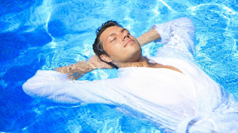 Junger Mann liegt auf Rücken im Pool, trägt weißen Bademantel
