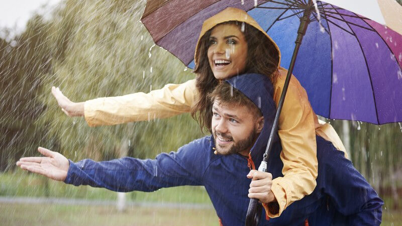 Die Hand ausstreckendes Paar in blauer und gelber Regenjacke unter einem bunten Schirm im Regen, er trägt sie Huckepack