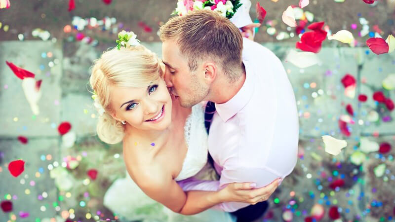 Rosenblätter fallen auf Hochzeitspaar herab, der Bräutigam küsst die blonde Braut