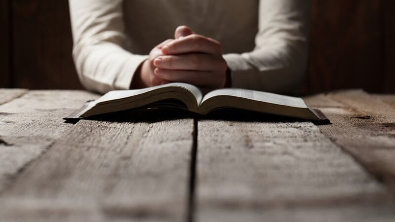 Zum Beten gefaltete Hände vor einer aufgeschlagenen Bibel auf altem Holztisch