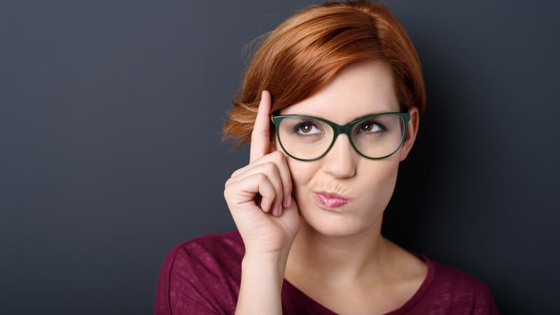 Frau mit rotbraunen Haaren hält nachdenkend den Zeigefinger neben ihre Brille und verzieht den Mund, grauer Hintergrund