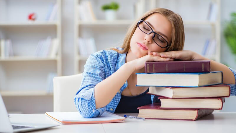 Junge Studentin in blauer Bluse und mit Brille sitzt am Schreibtisch und schläft auf ihren Büchern