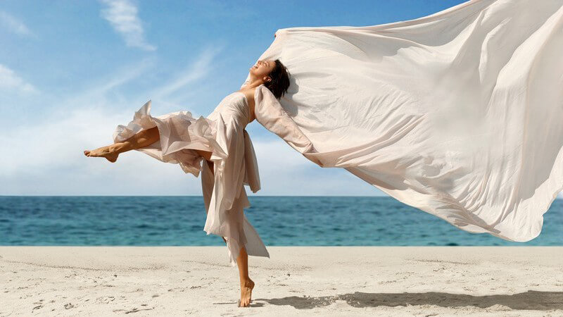Tanzende Frau mit beigem Kleid und großem, wehenden Tuch am Strand am Meer