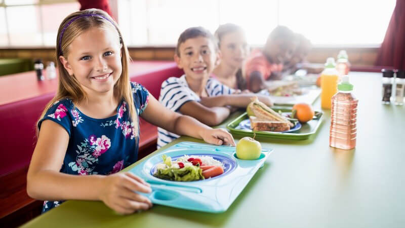 Mädchen in blauem Sommerkleid und andere Kinder sitzen mit ihrem Essen an einem großen grünen Tisch in der Schulkantine