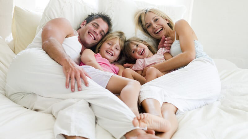 Junge Familie mit zwei Töchtern liegt zusammen im Bett und lacht