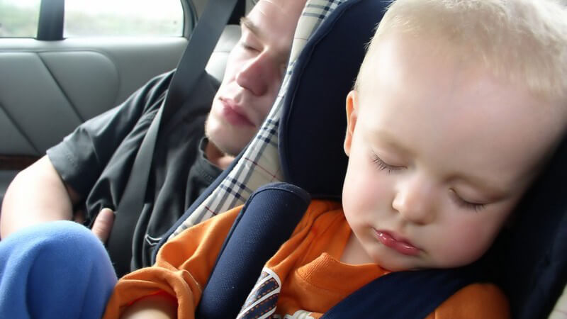Kind im Autokindersitz schläft, dahinter schläft junger Mann im Auto