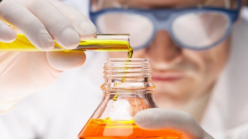 Laborarzt mit blauer Schutzbrille schüttet orange Flüssigkeit aus Reagenzglas in Flasche