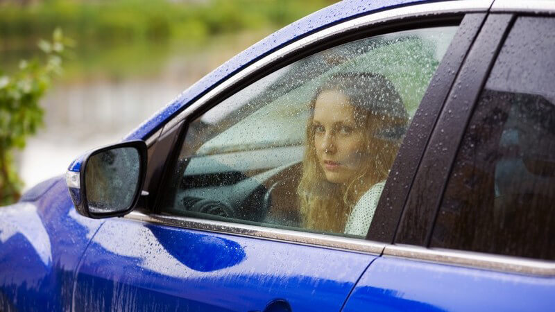 Junge Frau sitzt im blauen Auto und schaut aus Fenster, draußen regnet es