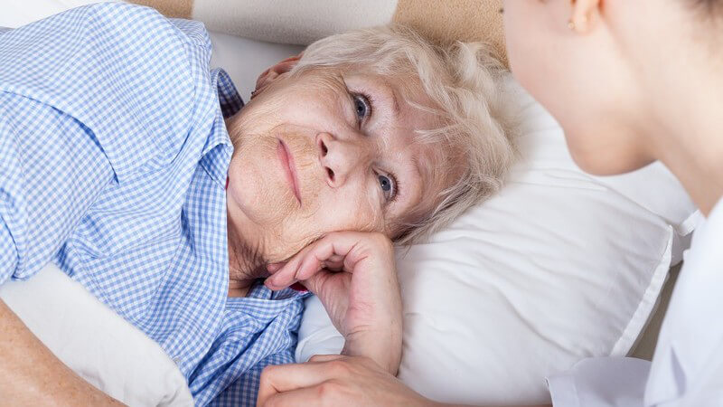 Ältere Frau liegt krank im Bett, betreut von Krankenschwester