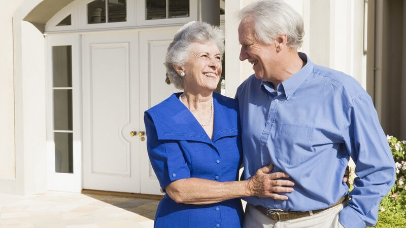 Senioren Paar steht lächelnd Arm in Arm vor Haus