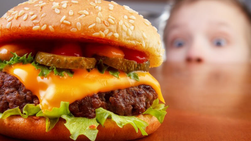 Dicker Burger auf einem Tisch, wird von einem Jungen im Hintergrund gierig beguckt