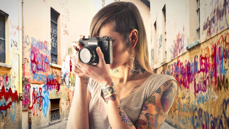 Tätowierte Fotografin steht in einer Gasse mit vielen Graffitis und guckt durch die Linse ihrer Kamera