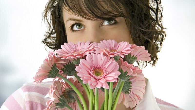 Junge Frau riecht an Strauß mit rosanen Blumen, Mund und Nase dahinter versteckt