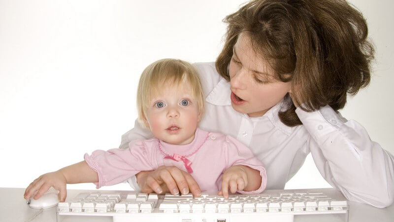 Mutter sitzt mit Kleinkind vor Computer, das Mädchen schaut neugierig auf Bildschirm