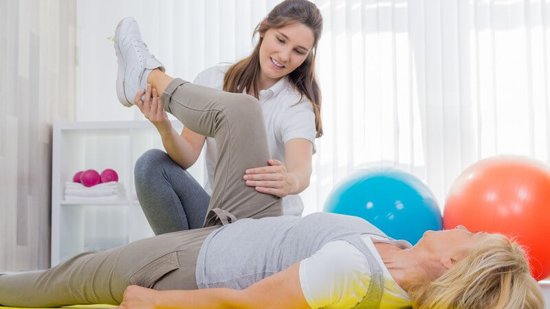 Physiotherapeutin macht eine Beinübung mit einer Seniorin auf gelber Gymnastikmatte