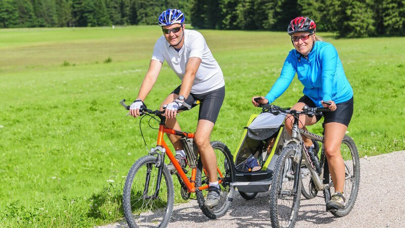 Elternpaar in Radsportbekleidung bei einer Radtour mit Mountainbikes, der Vater zieht einen Kinderfahrradanhänger