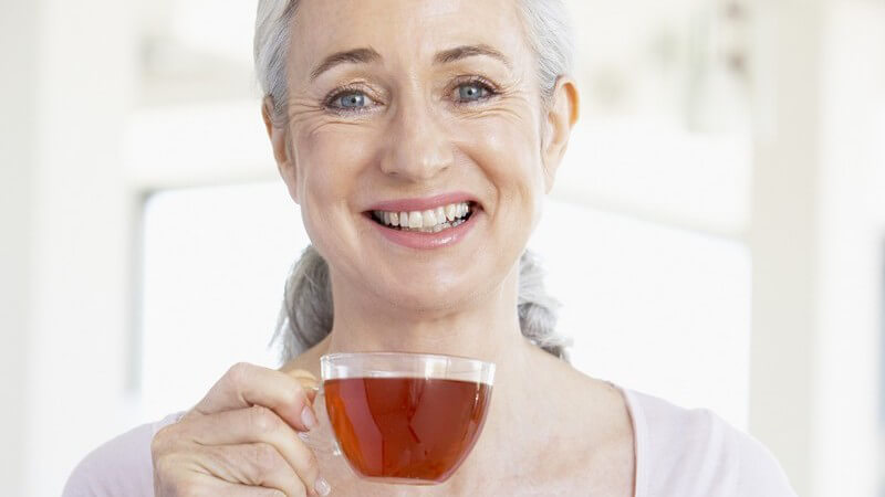 Ältere Frau hält Glas mit Tee in Hand und lacht in Kamera
