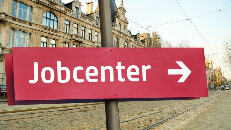 Roter Wegweiser mit der Aufschrift "Jobcenter" vor alten Gebäuden und Straßenbahngleisen