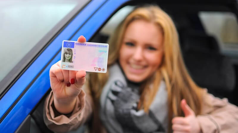 Junge Frau sitzt in blauem Auto und hält stolz ihren Führerschein aus dem Fenster