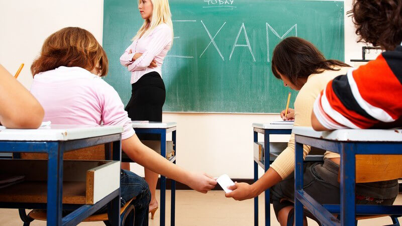 Rückenansicht Schulklasse bei Prüfung, zwei Schüler tauschen Zettel aus