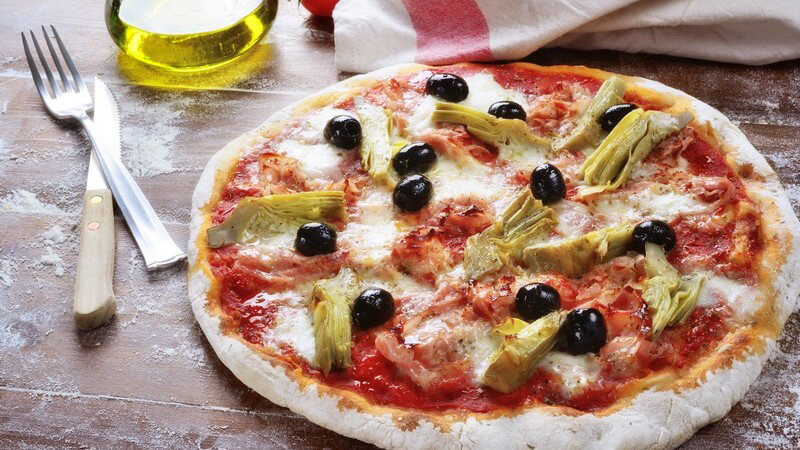 Pizza auf einem Holztisch voller Mehl, belegt mit schwarzen Oliven, Artischocken und Schinken