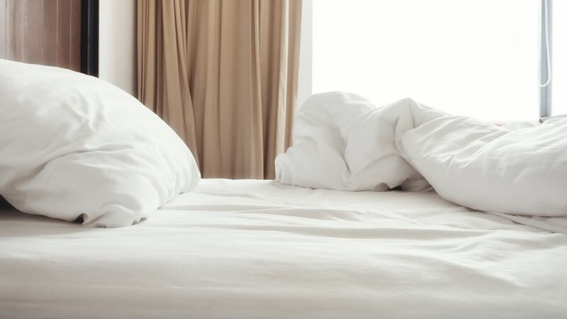 Ungemachtes Bett mit weißer Bettdecke und weißem Kopfkissen, braune Gardine im Hintergrund