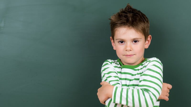 Junge in grün-weiß gestreiftem Longsleeve steht mit verschränkten Armen vor einer Tafel