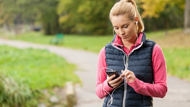 Junge Frau mit dunkelblauer Weste steht im Park und bedient ihren MP3-Player