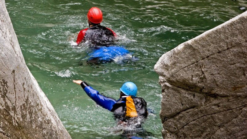 Zwei Menschen schwimmen in Neoprenanzügen in grünlichem Wasser zwischen Felsen, einer im roten und einer im blauen Helm