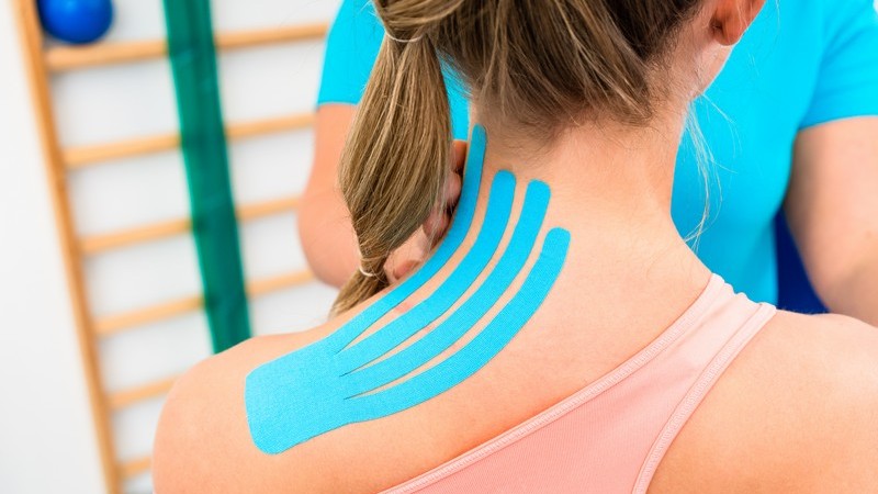 Junge Frau bekommt vier Streifen blaues Physio-Tape auf Schulter und Nacken angebracht