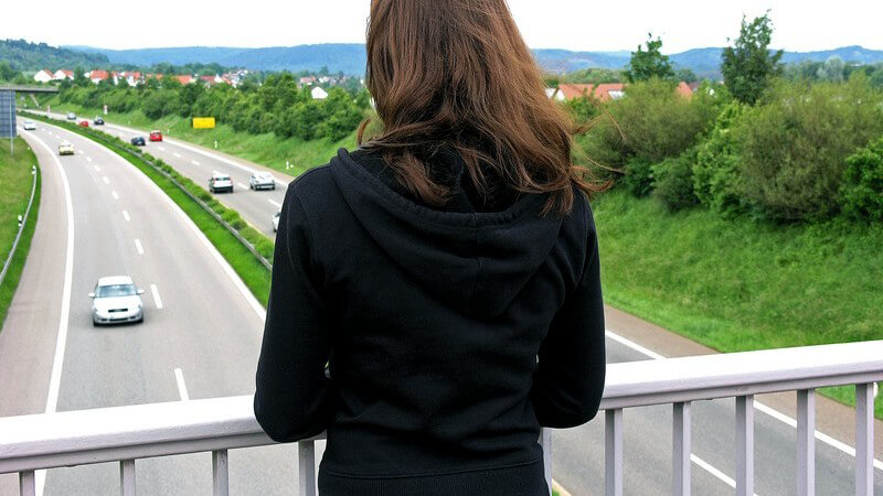 Brünettes Mädchen mit schwarzem Pulli blickt von einer Autobahnbrücke, Kamera von hinten