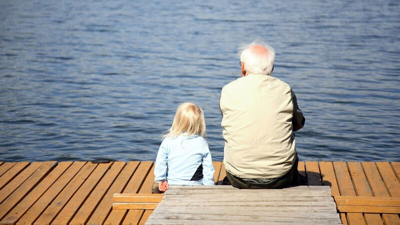 Rückenansicht Opa sitzt mit seiner Enkelin auf einem Steg am See und schaut aufs Wasser