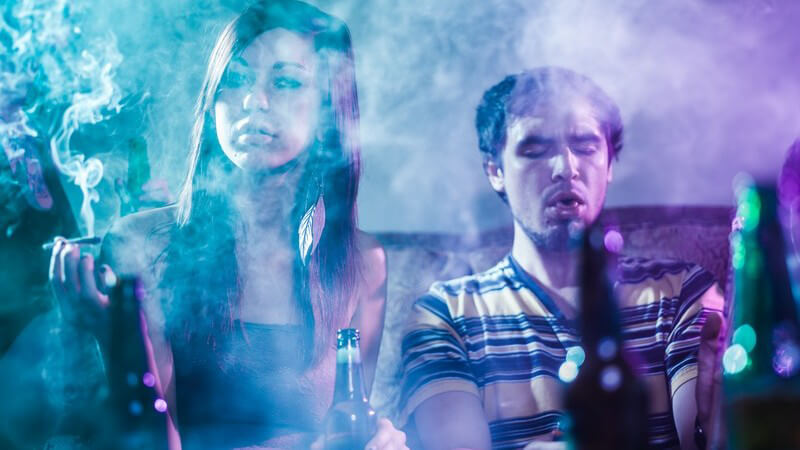 Jugendliche sitzen in einem vernebelten Raum, trinken Alkohol und rauchen Marihuana