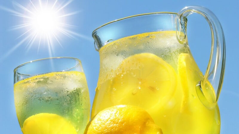 Zitronen vor Glaskanne und Glas mit kalter Zitronenlimonade unter blauem Himmel