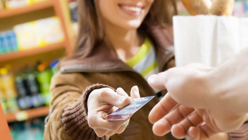 Junge Frau mit Einkaufstüte steht an Kasse und hält Kassierer Geldkarte hin
