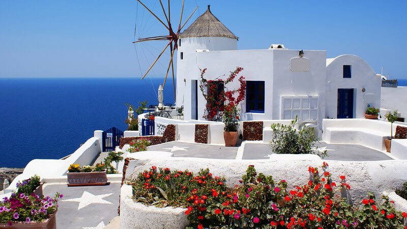 Kleine weiße Windmühle und rote Blumen vor strahlend blauem Himmel in Griechenland