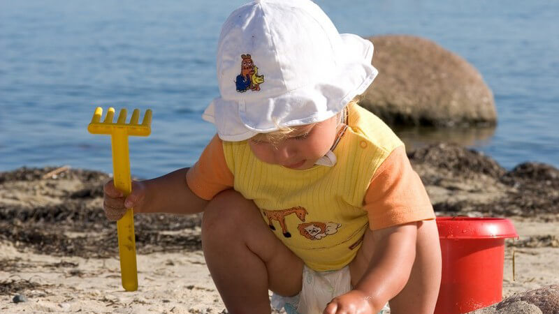 Kleinkind mit Windel und Hut hockt mit Schaufel neben Eimer am Strand