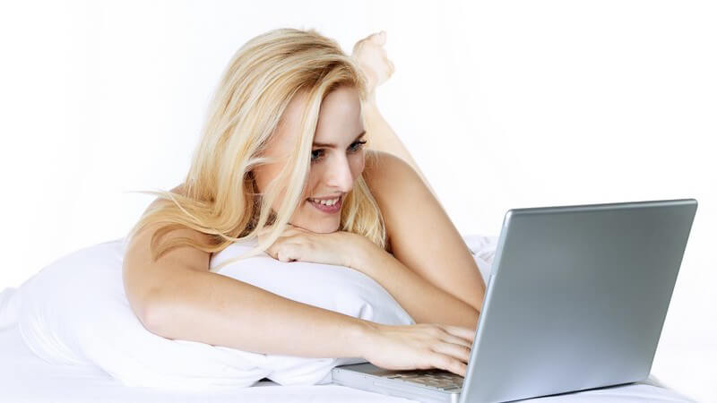 Blonde Frau liegt auf einem weißen Kissen und nutzt einen grauen Laptop