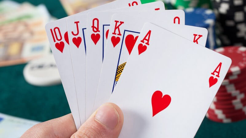 Hand hält fünf Pokerkarten mit einem "Royal Flush", im Hintergrund liegen Geldscheine und Poker-Chips
