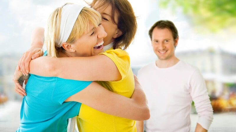 Zwei Freundinnen umarmen sich lachend, im Hintergrund lächelnder Mann