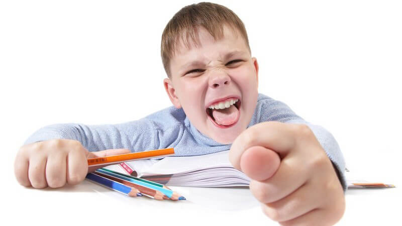 Kleiner Junge mit Buch und Buntstiften grinst mit offenem Mund frech in die Kamera