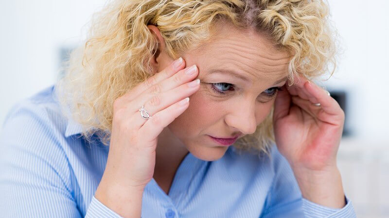 Frau mit Dauerwelle und blauer Bluse sitzt mit Kopfschmerzen im Büro und fasst sich an die Schläfen