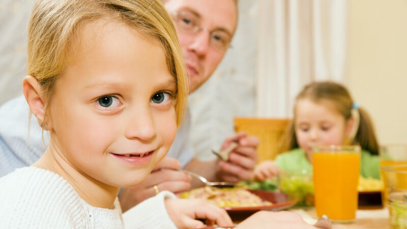 Familie am Tisch beim Essen, kleine Tochter lächelt schüchter in Kamera