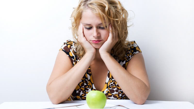 Frau sitzt unglücklich am Tisch vor Teller mit grünem Apfel zur Diät