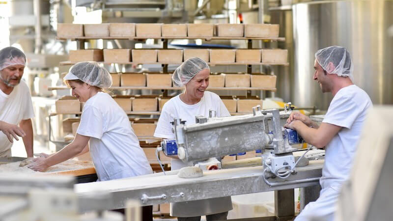 Vier Arbeiter/innen in weißer Arbeitskleidung und Haube in einer Großbäckerei